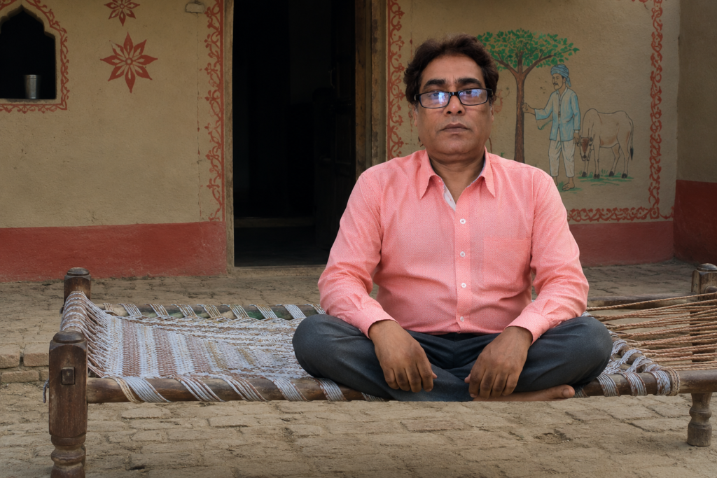 खाट की वापसी आधुनिक जीवनशैली के बीच परंपरा, स्वास्थ्य और पर्यावरण का पुनर्जागरण पारंपरिक भारतीय गांव के आंगन में लकड़ी की खाट पर बैठे चश्मा लगाए व्यक्ति की तस्वीर, जो देसी जीवनशैली, भारतीय संस्कृति और खाट की वापसी को दर्शाती है।