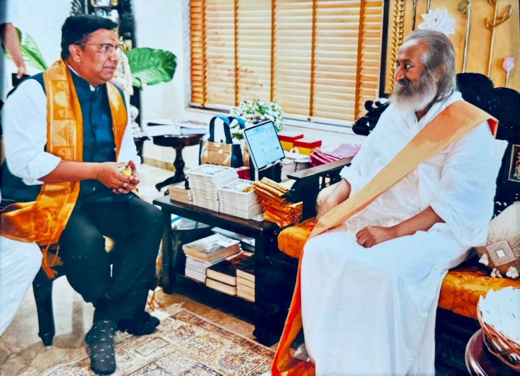 आध्यात्मिक गुरु Sri Sri Ravi Shankar अपने आश्रम में Prabhat Kumar से भेंट करते हुए। प्रभात कुमार पारंपरिक भगवा अंगवस्त्र पहने हुए बैठे हैं, जबकि श्री श्री रविशंकर सफेद वस्त्रों में मुस्कुराते हुए उनसे संवाद कर रहे हैं। मेज पर पुस्तकें रखी हैं, जो IIT पर आधारित आगामी पुस्तक के विमोचन और बौद्धिक चर्चा का प्रतीक हैं।